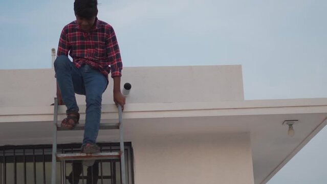 Indian male teenager descending from the roof of the house with the help of a metal staircase