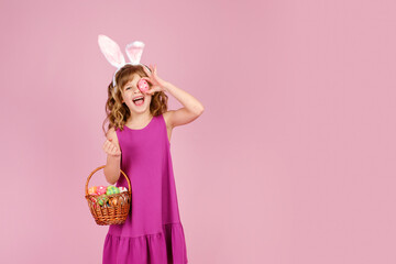Obraz na płótnie Canvas Happy child with long curly hair in fuchsia dress and bunny ears, smiling with open mouth and looking at camera while covering eye with Easter egg standing in pink studio with wicker basket