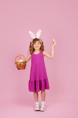 Obraz na płótnie Canvas Adorable cute girl with blond curly hair in stylish dress and bunny ears headband, happily smiling and looking at camera while demonstrating wicker basket and colorful Easter egg, vertical, copy space