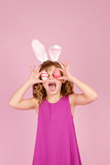 Adorable cheerful little girl with curly hair in fuchsia colored dress and bunny ear headband, smiling happily while covering eyes with eggs during Easter celebration, vertical orientation, copy space