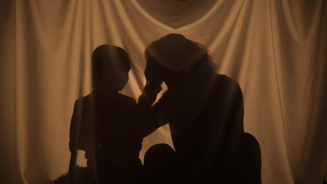 Silhouettes of woman and boy enjoying music with big headphones. Mom and son spend leisure together, sitting behind curtain at home. Puppet theater of shadows, play of shadow and light. Close up.