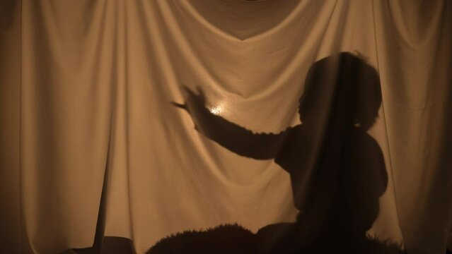 Silhouette of little boy dancing, gesturing hands and having fun. Child of primary age spend leisure, sitting behind curtain at home. Puppet theater of shadows, play of shadow and light. Close up.