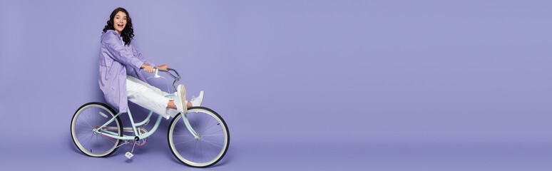 full length of amazed young woman in violet raincoat riding bicycle on purple, banner.