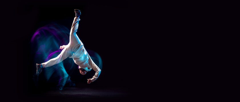 Hand stand. One energy young flexible sportive man dancing hip-hop or breakdance in white outfit on dark background in mixed blue neon light. Sport, art, action, moves