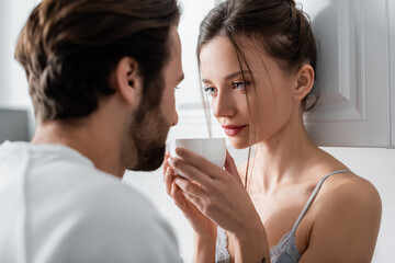 young woman in bra holding cup and looking at blurred bearded boyfriend.