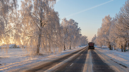 Tractor with trailer on a picturesque rural road at sunset. On the sides of the road there are beautiful trees in frost.