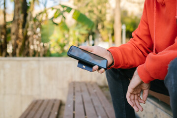 Young man wearing red hoodie holding phone and credit card in one hand. Sitting young man shopping outdoors.