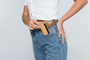 cropped view of young woman putting credit card with cashback in pocket on jeans isolated on grey.