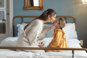 Tender mom kissing her baby girl with glitter on cheeks, playing at bedroom, sitting on bed with modern interior. The concept of a happy relationship between mother and daughter. Happy mothers day