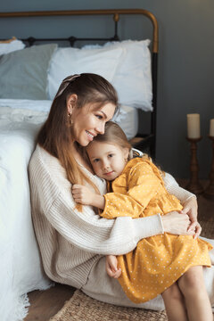 Vertical photo of happy young mother and daughter. Tender mom and baby girl sitting on floor and hugging at domestic living room with modern retro interior. Happy mothers day