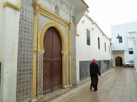 Moroccan woman in hijab walking past colorful entrance door to mosque in Medina of Rabat, Morocco.