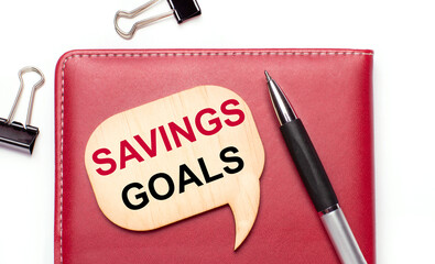 On a light background there are black paper clips, a pen, a burgundy notepad a wooden board with the text SAVINGS GOALS
