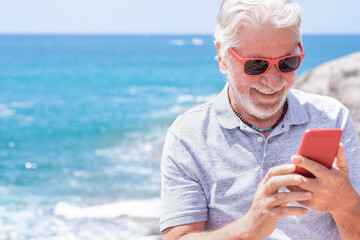 Smiling caucasian bearded grandfather in outdoors at sea enjoying sunny day and vacation. Senior man with red sunglasses using modern technologies and wireless connection. Horizon over water