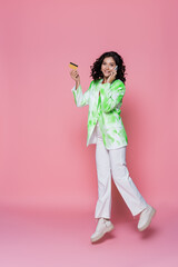 full length of smiling young woman in tie dye blazer holding credit card and talking on smartphone on pink.
