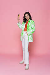 Fototapeta na wymiar full length of pleased young woman in tie-dye blazer holding credit card and posing with hand in pocket on pink.
