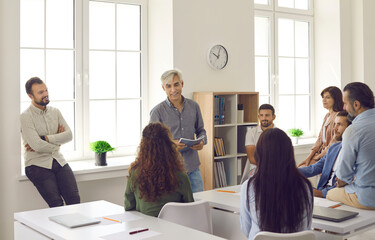 Team of people meeting for training session with mentor in modern office. Group of adult students or company employees sitting at desks in classroom and listening to experienced senior business coach - obrazy, fototapety, plakaty