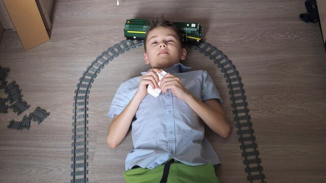 a teenager is playing with a train on the controls on the floor in the room. top view. toy railway. smiling, he turns his head to the moving locomotive. there is light from the window in the room