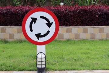 White and red roundabout sign on the road with plants in park in the village background. Sign and...
