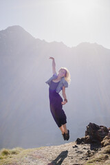 A young beautiful girl in a blue long dress and a denim jacket jumps posing against the backdrop of the high Kazbek mountains in the rays of the dawn sun. Georgia. Europe