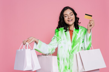 cheerful young woman in tie dye blazer holding credit card and shopping bags isolated on pink.
