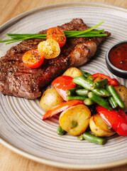 Grilled steak with vegetables and sauce. Juicy steak with tomatoes, potatoes, peppers and green beans in a plate on a wooden background. Delicious hearty lunch. Meat with baked vegetables. Close up.