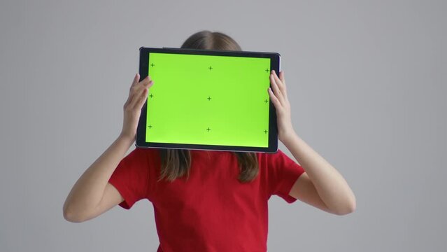 A blonde teenager girl in a red T-shirt stands with a tablet in her hands in front of her head with a green screen against a white wall