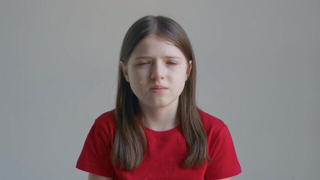 A blonde teenager girl in a red T-shirt cries and wipes her tears with her hands against the background of a white wall