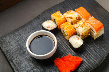 Assorted rolls on a black board next to soy sauce and ginger on a dark background.