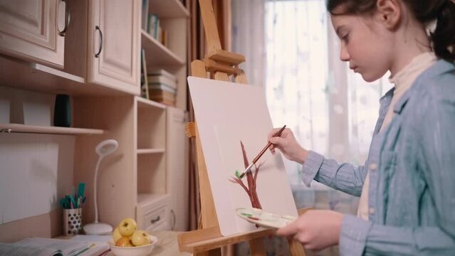 Teenager girl paints tree on paper at home. Slider medium shot.