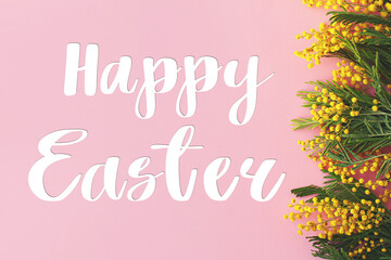 Happy Easter text on mimosa flowers flat lay on pink background. Stylish seasons greeting card. Handwritten lettering happy Easter