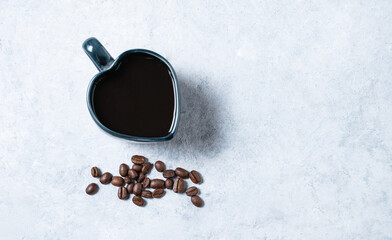 A cup of black espresso coffee in the shape of a heart with scattered grains on a blue background. Top view and copy space
