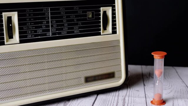 Old Vintage Receiver Standing on a Table, with an Hourglass Counting Down the Time. Sand pours inside a glass flask, counting the time of the end of the era of the 70s, 80s, 90s. Outdated electronics.