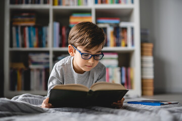 One caucasian boy lying on the floor at home in day reading a book front view wearing eyeglasses copy space real people - obrazy, fototapety, plakaty