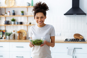 Healthy diet eating. African American young female holding bowl of fresh salad standing in the home...