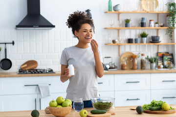 Happy african american woman standing at the cuisine table in the home kitchen drinking dietary supplements, looking away and smiling friendly, healthy lifestyle concept