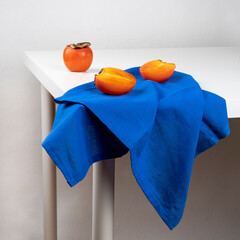Still life with persimmon fruits and a blue linen towel lying on a white table.