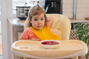 A small child eats vegetable soup with a spoon while sitting on a high chair