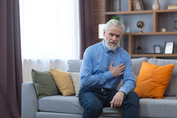 Older single man alone at home, sitting on sofa, holding hands on chest, has severe chest pain