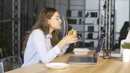 Young Businesswoman Eating Hamburger while Working on Laptop at the Office Table