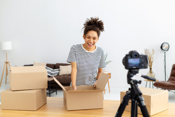 Fototapeta na wymiar African American female blogger vlogger freelancer using camera to recording video content for his followers, makes an overview of the products, goods. Young woman unpacking boxes on camera