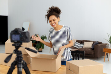 Plakat African American female blogger vlogger freelancer using camera to recording video content for his followers, makes an overview of the products, goods. Young woman unpacking boxes on camera