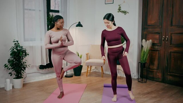 African american woman yoga instructor teaching asian lady newcomer to practice tree asana, exercising together at home