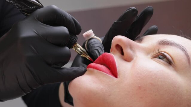 the process of applying permanent makeup tattoo red color on the lips of a woman. Specialist applying paint with tattoo tool