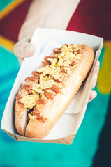 Freshly prepared hotdog with salsa and fried onions in a paper box in a hand. Food delivery concept. Green background.