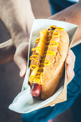 Freshly prepared cheesy hotdog in a paper box in a hand. Food delivery or lyfestyle concept.