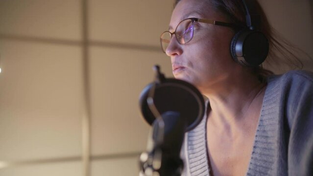 Middle aged woman blogger in headphones broadcasting her speech using microphone with pop filter. Modern audio sound studio recording technology concept image.