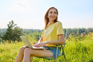 Naklejka na ściany i meble Summer vacation in wild meadow, adult woman resting on folding chair for camping using laptop