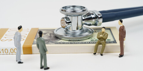 On a white surface lies a pack of dollars, with a stethoscope on the dollars. Nearby are figures of businessmen.