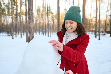 Cheerful African American woman making a snowman in a snow covered woodland. Beautiful sunbeams falling on the snowy forest path. Enjoy wonderful winter, leisure games and activities outdoor