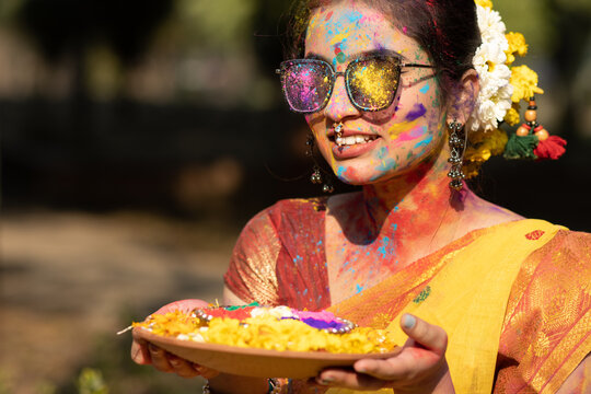 Holi festival of color, young beautiful girl or woman or lady in indian attire saree playing holi with dry organic color or colour or gulal or abeer or Holi powder placed in clay bowl with flowers